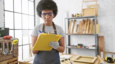 Kıvırcık saçlı, güvenlik gözlüğü takan ve kot önlüklü gülümseyen zenci bir marangoz parlak, düzenli bir ahşap atölyesinde panoya yazı yazıyor..