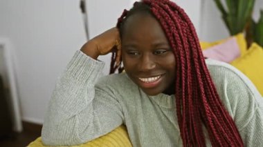 Kapalı bir ortamda gülümseyen kırmızı örgülü Afrikalı bir kadın. Mutluluk saçıyor ve sıradan bir tarz sergiliyor..