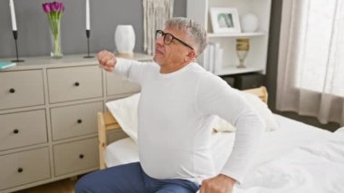 Uykulu bir sabah esnemesi, gri saçlı, orta yaşlı bir adam tamamen esniyor ve pijamalarıyla esniyor, yatak odasında rahat bir başlangıç yapmak için erken kalkıyor.