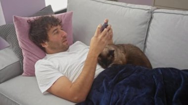 Evdeki kanepede uzanmış genç bir İspanyol adam yanında siyam kedisi olan akıllı bir telefonla mavi bir battaniyenin altında yatıyor..