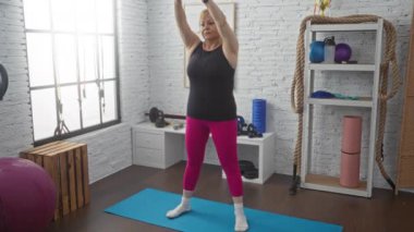 Yaşlı bir kadın spor salonunda esneme egzersizleri yapıyor. Pembe taytlar ve siyah bir bluzla, halatlar, mavi bir paspas, ağırlıklar ve büyük bir pencere eşliğinde..