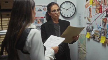 İki kadın müfettiş bir ofiste kanıt panosu, belgeler, saat ve dosya dolaplarıyla bir davayı tartışıyor..