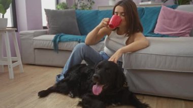 Genç bir kadın, rahat bir oturma odasında siyah labrador köpeğini okşarken bir fincan kahveyle rahatlıyor, modern mobilyalar ve canlı dekorlar sunuyor..