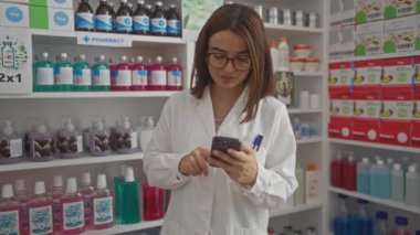Gözlüklü ve beyaz önlüklü genç bir İspanyol kadın bir eczanede duruyor, elinde cep telefonu var, raflarda çeşitli ilaçlar ve sağlık ürünleri var..