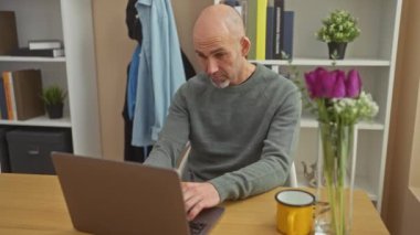 Kel ve sakallı bir adam, modern bir ofis ortamında dizüstü bilgisayar kullanırken çeşitli duygular ifade eder..