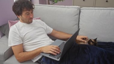 Genç bir adam evdeki kanepede dizüstü bilgisayarda çalışıyor mavi bir battaniyenin altında bir kediyi okşuyor..