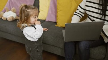 Konforlu bir oturma odasında, odaklanmış bir kadın, meraklı kızının yanında tulum giyerek dizüstü bilgisayarda çalışır..