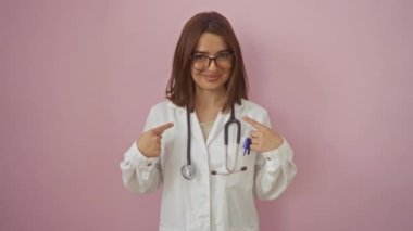 Doktor üniforması giyen genç İspanyol kadın mutlu bir şekilde el ve parmaklarıyla izole edilmiş pembe arka planı işaret ediyor.