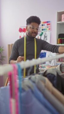 Şerit mezuralı Afrikalı Amerikalı terzi elbise raflarının arasında renkli bir atölyede çalışıyor..