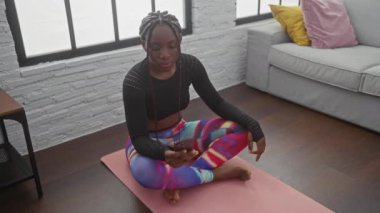 Modern bir oturma odasında akıllı telefonla yoga yapan Afrikalı Amerikalı bir kadın.
