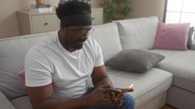 Sakallı Afrikalı genç bir Amerikalı oturma odasındaki koltukta akıllı telefon kullanarak rahat ve odaklanmış bir ortam yaratıyor..