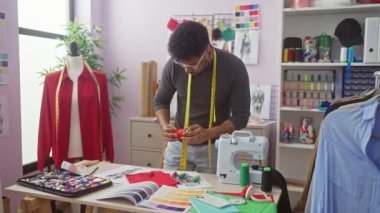 Afrikalı Amerikalı erkek terzi renkli bir atölyede, manken ve dikiş makinesiyle mezura kullanıyor.