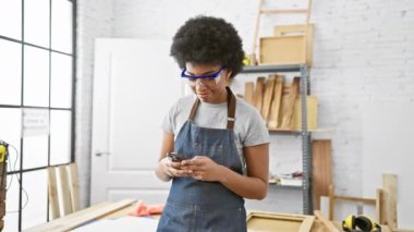 Marangozluk atölyesinde güvenlik gözlüğü ve önlükle telefon kullanan Afrikalı Amerikalı bir kadın.