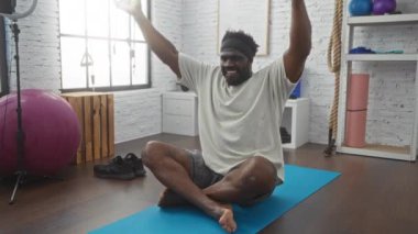 Gülümseyen, genç, Afro-Amerikalı bir adam, spor malzemeleriyle dolu parlak bir spor salonunda mavi yoga minderinin üzerinde egzersiz yapıyor..