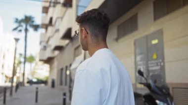Beyaz tişört ve gözlük takmış, şehir arka planında yüksek binalar ve palmiye ağaçları olan yakışıklı İspanyol bir adam güneşli bir günde şehir sokaklarında dikiliyor..