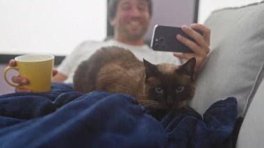 Gülümseyen bir adam, sıcak bir odada sarı bir kupa tutarken mavi bir battaniyenin üzerinde siyam kedisiyle selfie çeker..