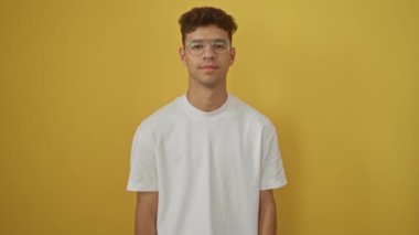 Beyaz tişört giyen gülümseyen genç İspanyol adam, kendinden emin ve mutlu duruyor, izole edilmiş sarı arka planda dişlerini gösteriyor.