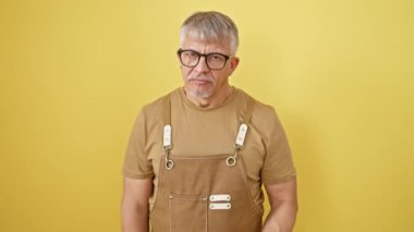 Dikkat et! Orta yaşlı, gri saçlı, önlüklü ve gözlüklü bir adam gözünü işaret ederek sarı bir zemin üzerinde duruyor.