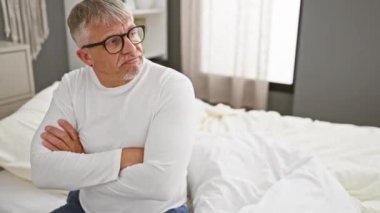 Kendine güveni tam, beyaz saçlı, orta yaşlı pijamalı, yana odaklanmış, kollarını kavuşturmuş, yatak odasında pozitiflik saçan bir adam.