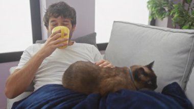 A young man sitting on a sofa in a cozy living room, relaxing with his siamese cat while holding a yellow cup.