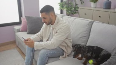 Oturma odasında cep telefonu kullanan genç bir İspanyol, tekel köpeğinin yanındaki koltukta otururken, rahat ve modern bir ev atmosferi yaratıyor..