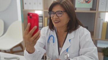 Kısa saçlı, beyaz önlüklü ve gözlüklü bir kadın doktor klinikteki bir video görüşmesi sırasında elinde kırmızı bir telefon tutuyor..