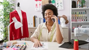 Gülümseyen Afrikalı bir kadın moda tasarımcısı atölyesinde konuşuyor. Etrafı çizimler ve dikiş malzemeleriyle çevrili..