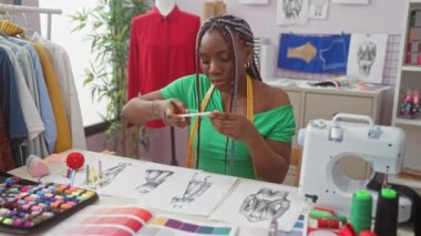 Renkli iplikler ve tekstil ürünleriyle çevrili bir terzide moda tasarımlarını analiz eden Afrikalı Amerikalı kadın..