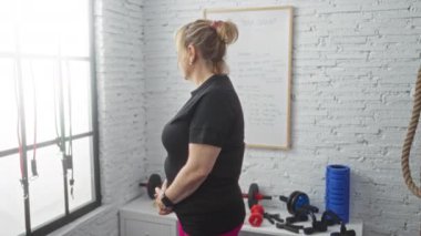 Siyah spor giyim giymiş sarışın bir kadın spor kıyafetleri içinde kollarını bağlar ve beyaz tuğladan duvarlar ve beyaz tahtaya çizilmiş spor kıyafetleri giyer..