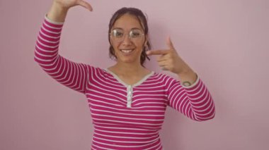 Işıl ışıl genç İspanyol kadın pembe izole arka plan üzerinden el çerçevesi yapıyor, çizgili tişört giyiyor, fotoğraf konseptini yeniliyor.