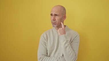 Sarı kazak giyen dalgın kel adam, derin düşüncelere dalmış, eli çenesinde, dikkatle, izole edilmiş arka plana bakıyor.