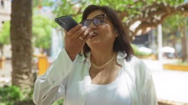 Kısa saçlı, cep telefonu kullanan ve güneşli bir günde şehir parkında gülümseyen yaşlı bir kadın..