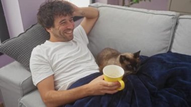 Genç bir İspanyol adam evdeki kanepede bir fincan kahveyle dinleniyor, oturma odasında siyam kedisiyle eğleniyor..