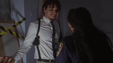 Dedektifler olarak tasvir edilen bir erkek ve kadın, yoğun bir şekilde aydınlatılmış bir odada suç mahalli bandıyla kordon altına alınmış bir şekilde sohbet ediyorlar..