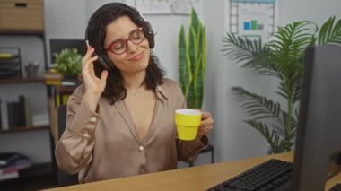 Genç bir İspanyol kadın şık bir ofiste elinde fincanla dans ediyor, kulaklıkla müzik dinliyor ve gözleri kapalı gülümsüyor..