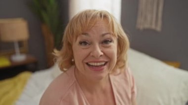 Orta yaşlı, sarışın bir kadın kameraya el sallıyor ve gülümsüyor yumuşak ışıklandırmalı ve sade dekorlu bir yatak odasında rahat bir yatakta otururken..