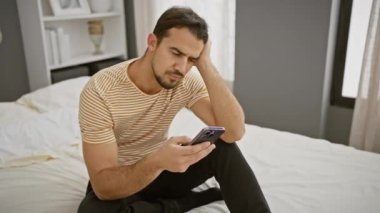 Düşünceli, sakallı İspanyol bir adam modern bir yatak odasında bir yatağa oturmuş akıllı telefonuna bakıyor..