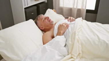 Gri saçlı adam güneşli bir yatak odasında uyanıyor.