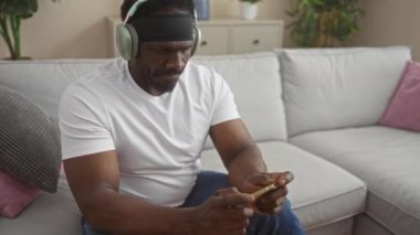 Oturma odasında oturan Afro-Amerikalı genç bir adam telefonuyla video oyunu oynuyor kulaklık takıyor, rahat bir koltukta oturuyor..