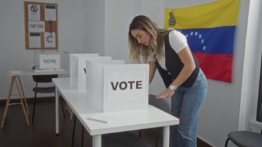 Venezuela 'da bir seçim koleji odasında genç sarışın bir kadın. Arka planda Venezuela bayrağı olan bir oy kabininin yanında duruyor..