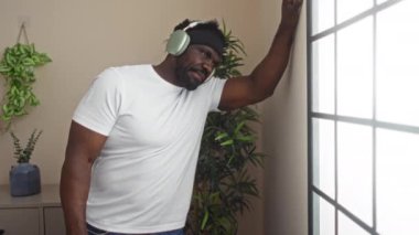 Kulaklık takan yakışıklı Afro-Amerikalı bir adam ev bitkileri ve ev dekorlarıyla çevrili, rahat bir oturma odasında duvara yaslanıyor..