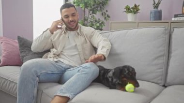 Gülümseyen İspanyol bir adam, rahat bir oturma odasındaki kanepede tekel okşarken telefonla konuşuyor..