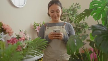 Çiçekçisine mutluluk ve güven saçan, akıllı telefonunu kullanırken dişleriyle gülümseyen parlak, genç İspanyol kadın çiçekçi.