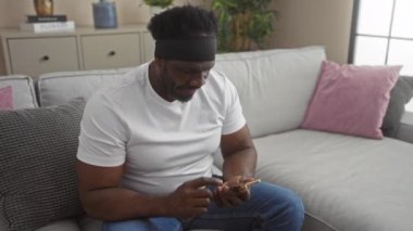 Genç, yakışıklı, sakallı Afrikalı Amerikalı bir adam rahat oturma odasında rahat bir kanepede otururken akıllı telefonuyla iletişim kuruyor..