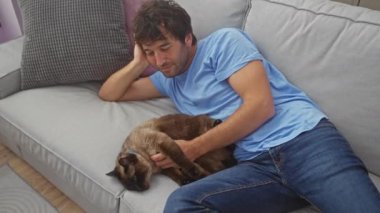 Mavi gömlekli rahat bir genç adam, içeride gri bir kanepede siyam kedisiyle sarılıyor..