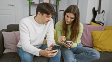 Bir erkek ve bir kadın, arka planda gitarlı, rahat bir oturma odasında, akıllı telefonlarına odaklanmış bir şekilde bir koltuğa yakın otururlar..