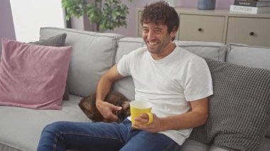 Beyaz gömlekli ve kot pantolonlu genç bir İspanyol bir adam oturma odasında oturuyor, gülümsüyor ve sıcak bir içeceğin tadını çıkarırken gri bir kanepede bir kediyi okşuyor..