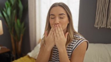 Çekici genç bir kadın, sıcak bir yatak odasında oturuyor, yüzünde mutlu bir ifadeyle bir öpücük üflüyor, yumuşak ışık ve modern dekorla çevrili..