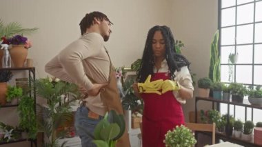 Bir kadın ve bir erkek, çeşitli saksı bitkileri ve çiçeklerle çevrili canlı bir çiçekçide neşeli bir sohbete girişiyor..