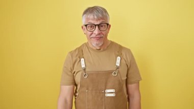 Neşeli orta yaşlı bir adam, gri saçlı ve gözlüklü, önlüklü, el hareketleriyle karşılıyor. Sarı bir arka planda izole edilmiş. Onun davetkar gülümsemesi seni buraya çağırıyor..